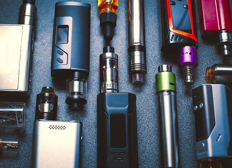 The image shows a variety of vape devices and e-cigarettes arranged on a dark surface. The devices come in different shapes, sizes, and colors, featuring a mix of metallic and plastic components. Many of them have visible tanks for e-liquid and some have digital displays and buttons for adjusting settings.