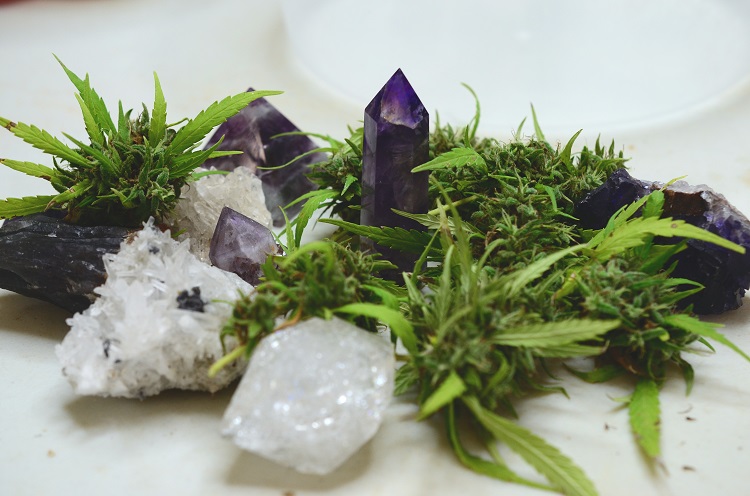 A mix of clear and purple crystals with green cannabis buds and leaves arranged on a flat surface.