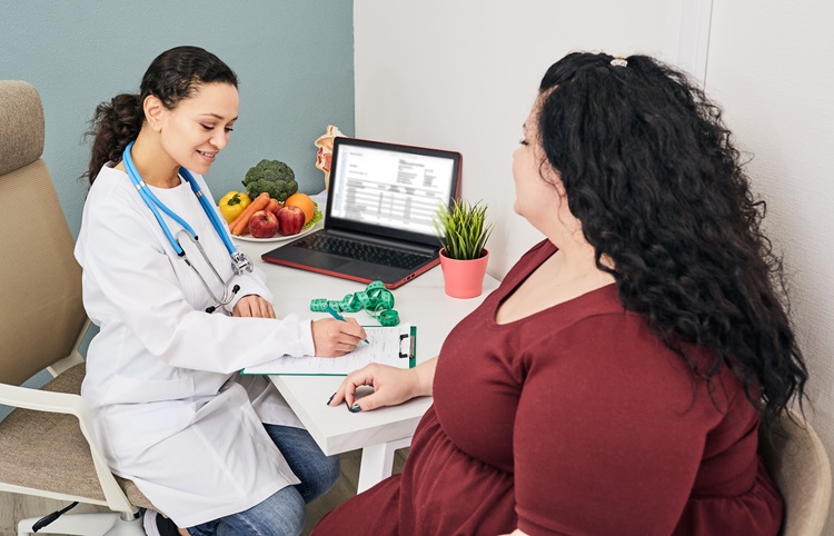 La imagen muestra a un profesional de la salud consultando con un paciente en un consultorio. El trabajador de la salud, con una bata blanca y un estetoscopio, escribe en un portapapeles. El paciente, sentado enfrente con un vestido granate, participa en la conversación. Una computadora portátil, un plato de verduras y una cinta métrica están sobre la mesa, lo que sugiere una discusión sobre salud o nutrición.