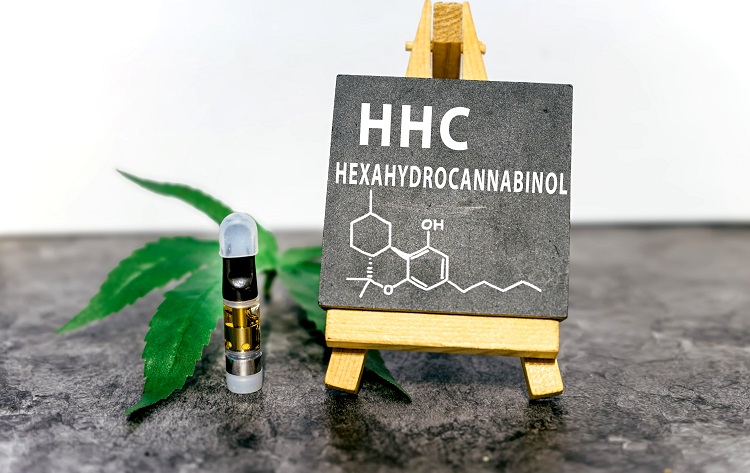 Slika prikazuje majhno tablo na stojalu s kemično strukturo in besedilom "HHC HEXAHYDROCANNABINOL". Poleg table je list konoplje in vape kartuša, ki vsebuje zlato obarvano tekočino. Ozadje je navadna bela stena, površina pa je temen, teksturiran material.