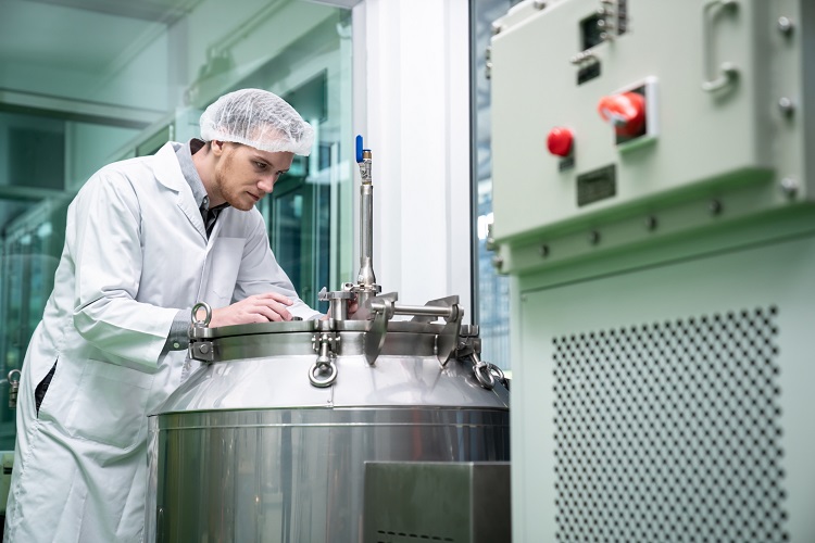 A imagem mostra uma pessoa com um jaleco branco e uma rede de cabelo, provavelmente um técnico ou cientista, trabalhando com um grande tanque de aço inoxidável ou vaso industrial. O indivíduo parece estar focado, possivelmente monitorando ou ajustando o equipamento. O cenário sugere um laboratório ou um ambiente de sala limpa, comumente encontrado em indústrias como farmacêutica, biotecnologia ou processamento de alimentos. Ao fundo, há também alguns equipamentos do painel de controle visíveis.